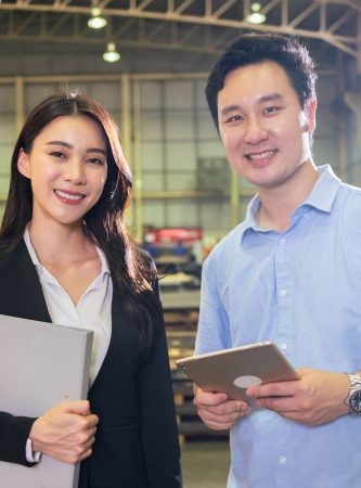 Portrait of Asian business man and woman work in manufacturing plant. Attractive factory engineer people in formalwear processes product at manufactory warehouse then looking at camera with happiness.